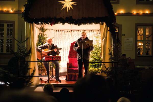 Zwei Männer auf einer Bühne in weihnachtlichem Ambiente und mit einer Gitarre und einem Liederheft © Henry Tornow Zwei Männer auf einer Bühne in weihnachtlichem Ambiente und mit einer Gitarre und einem Liederheft © Henry Tornow